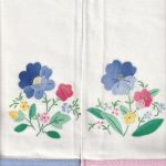 Set of 2 Hand-Stitched Appliqué Guest Towels in white, each with colorful embroidered flowers and green leaves. One has a blue hemstitch pastel border, the other pink; both share identical floral designs.