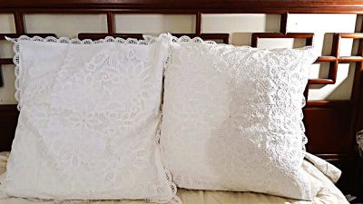 Two white decorative pillows with intricate lace patterns rest side by side against a wooden headboard on a light-colored bed.