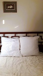 Two large white lace pillows rest against a wooden headboard with a geometric pattern. A framed picture hangs on the wall above the bed, and sunlight casts a small bright spot on the cream wall.