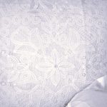 A close-up of a white lace pillow with intricate floral and circular patterns, partially covered at the bottom corner by a white fabric.