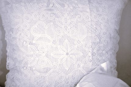 A close-up of a white lace pillow with intricate floral and circular patterns, partially covered at the bottom corner by a white fabric.