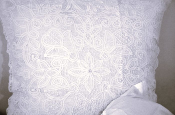 A close-up of a white lace pillow with intricate floral and circular patterns, partially covered at the bottom corner by a white fabric.