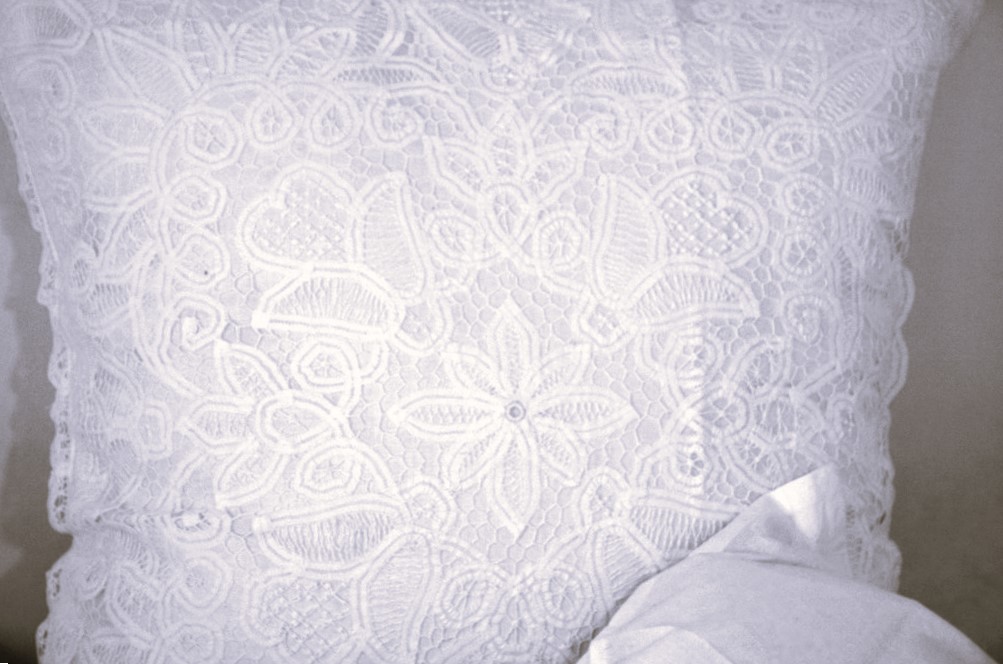 White Solid Battenbutg Euro 26sq IMG3053 amended A close-up of a white lace pillow with intricate floral and circular patterns, partially covered at the bottom corner by a white fabric.