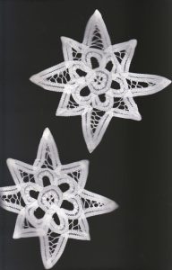 Two "Wish Upon a Star" handmade white Battenburg lace Christmas ornaments, each a 6-point star doily with delicate stitching and open spaces, are displayed on a black background. Part of the Trousseau Heritage Lace collection.