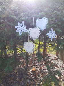 A Wish Upon a Star—Handmade White Battenburg Lace Christmas Ornament hangs from a ribbon on a green pine tree outdoors as sunlight streams through branches and fallen leaves cover the ground below.