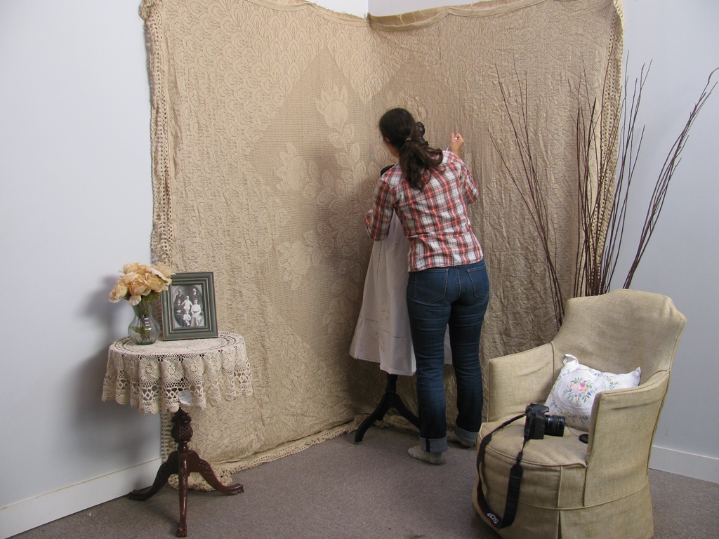 A woman in a plaid shirt arranges a dress on a mannequin before the Peacock Lace Photography Backdrop – Hand Loom Cotton. Nearby are a table with flowers and photo frame, and an armchair with a camera on it.