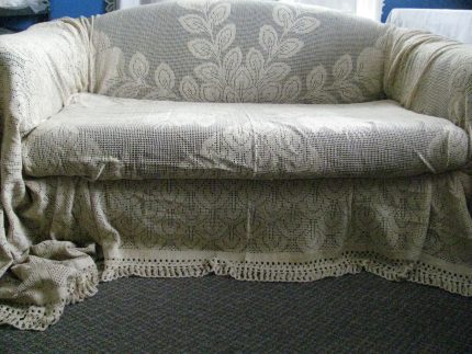 The Peacock Lace Sofa & Slip Cover, featuring a beige crocheted leafy pattern with fringed edges, is draped over a sofa placed on a gray carpet in a softly lit room.