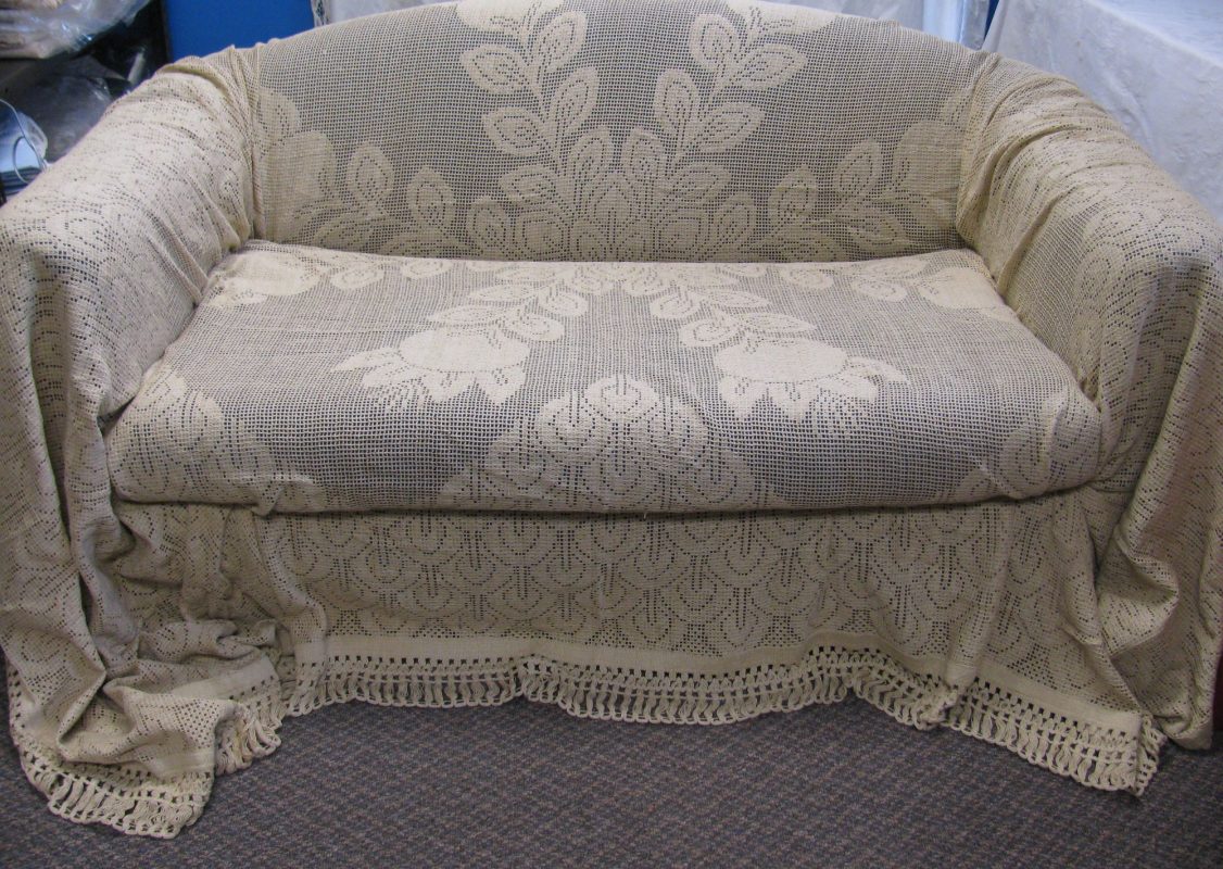 A beige crochet lace cover drapes over a small upholstered sofa with rounded arms. The cover features floral and geometric patterns and extends down to the floor, partially pooling on a gray carpet.