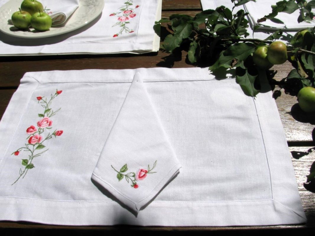 The American Beauty Embroidered Roses Outdoor Dining Entertaining Set, featuring white linen placemats and napkins with pink rose embroidery, is displayed on a wooden table with green apples and sunlight casting soft shadows.
