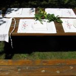 An outdoor wooden picnic table is set with the American Beauty Embroidered Roses Outdoor Dining Entertaining Set—4 placemats and 4 napkins—accented by green apples and leafy branches, in sunlight and shade.
