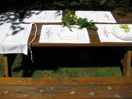 An outdoor wooden picnic table is set with the American Beauty Embroidered Roses Outdoor Dining Entertaining Set—4 placemats and 4 napkins—accented by green apples and leafy branches, in sunlight and shade.