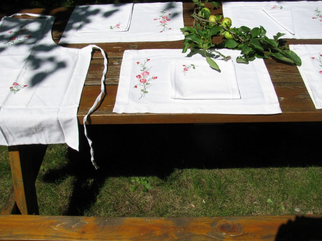 A wooden outdoor table is set with the American Beauty Embroidered Roses Outdoor Dining Entertaining Set—4 placemats and 4 napkins, each adorned with small pink flowers—plus green apples and leaves in sunlight.