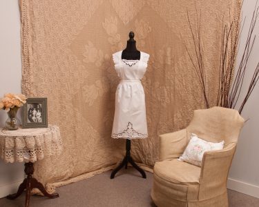 A vintage-style room with a beige patterned backdrop, a mannequin dressed in the Victorian Whitework Rose Apron – Cutwork Pinafore, a round lace-covered table with flowers and a framed photo, a beige floral armchair, and tall decorative branches.
