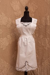 The Victorian Whitework Rose Apron – Cutwork Pinafore, featuring lace trim and embroidered details, is shown on a black dress form against a beige floral-patterned fabric background.