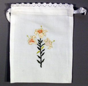 A white drawstring pouch with a crocheted lace trim at the top, featuring an embroidered design of three light-colored lilies with green stems and leaves on the front.