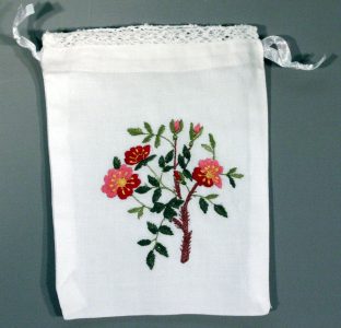 A white fabric pouch with lace trim and ribbon drawstrings, featuring embroidered Alberta Provincial Flower Wild Rose design.
