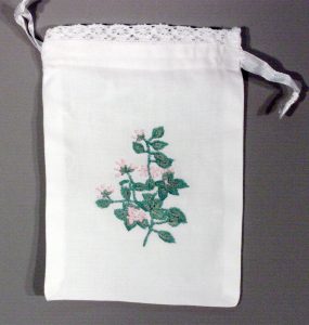 A white fabric drawstring bag with a lace trim at the top and an embroidered design of pink flowers with green leaves in the center.