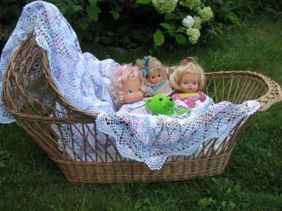 Three baby dolls and a green stuffed frog sit in a wicker bassinet lined with a white lace blanket, set on grass with green plants and white flowers in the background.