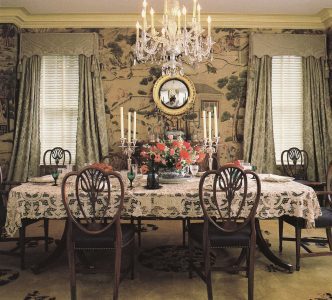 Elegant dining room with lace-covered table set for a meal, surrounded by wooden chairs. Crystal chandelier, floral centerpiece, and candelabras decorate the table. Large windows with drapes and ornate wallpaper complete the classic look.